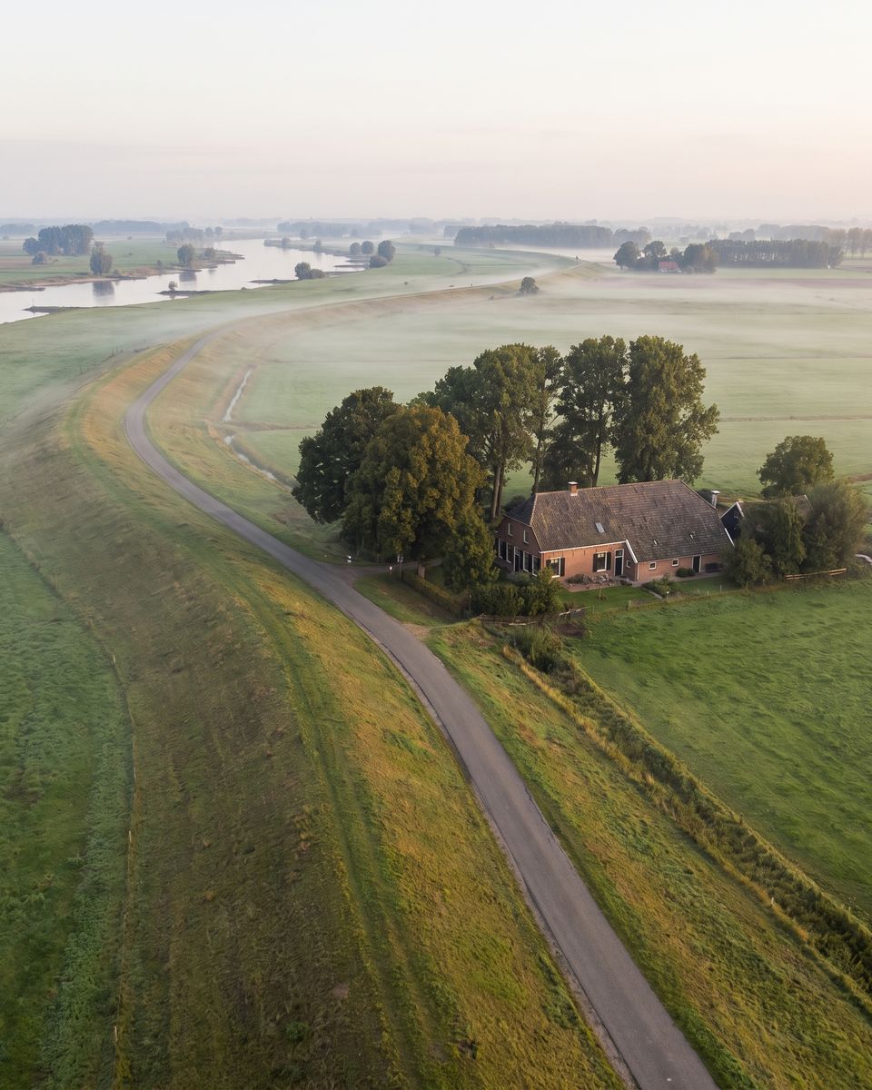 Dijk langs de IJssel met een boerderij tussen de bomen in de ochtendnevel