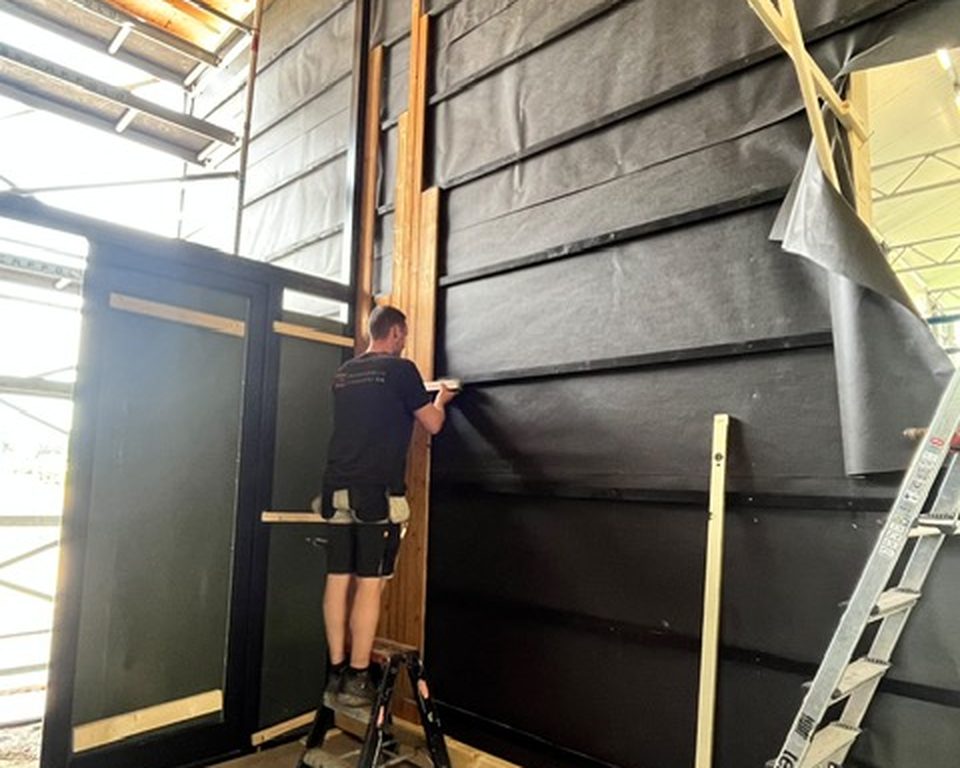 Collega van Groters werkt aan een houten gevelvlak met zwarte bekleding, binnen in een gebouw met steiger