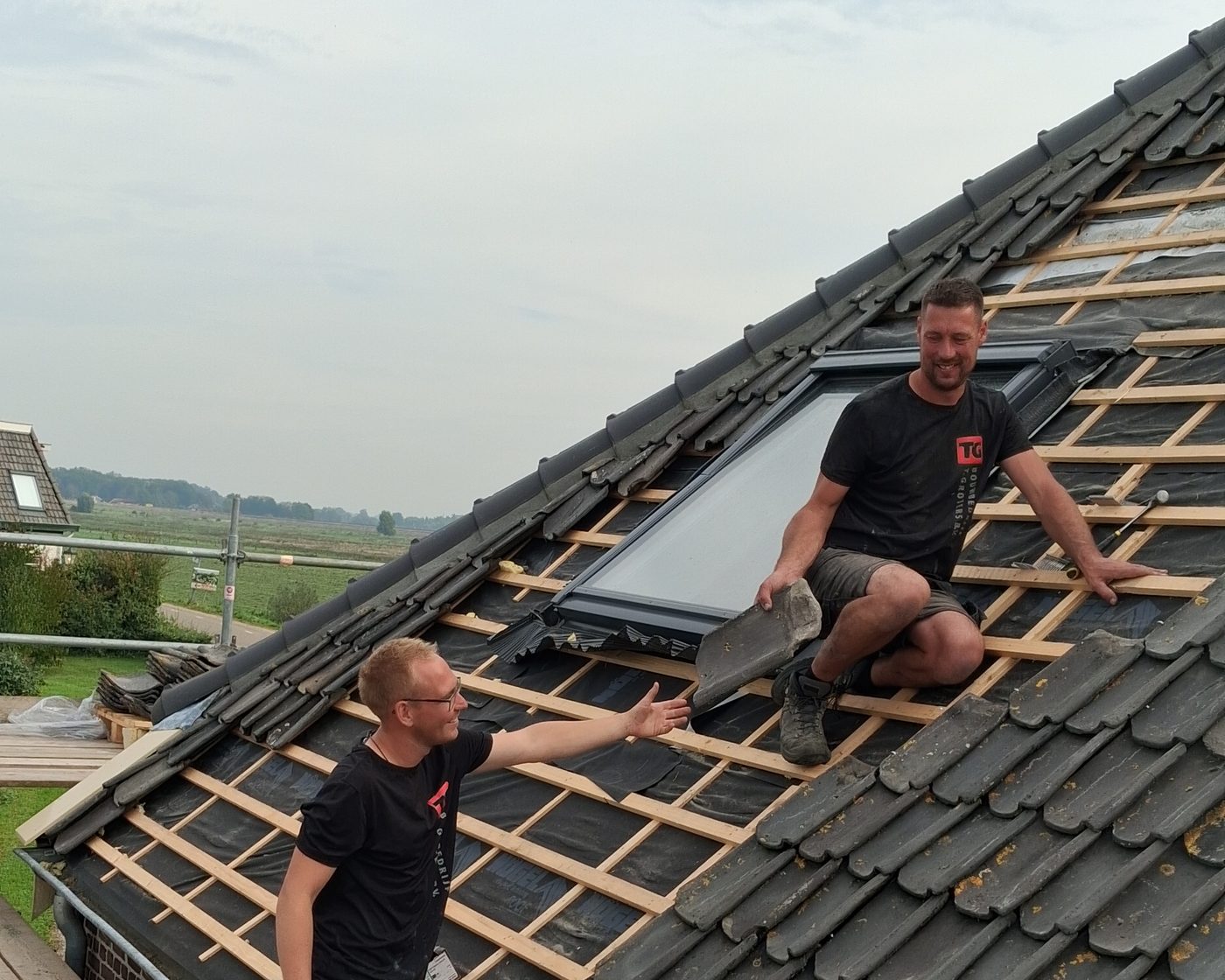 Twee collega's van Groters aan het werk op een dak, met een dakraam en dakpannen in de hand
